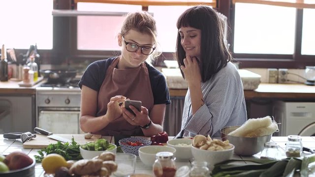 Two attractive hipster girls in casual clothing relax and chat over kitchen counter while getting ready to cook big festive meal for their family and friends from organic, fresh biologic ingredients