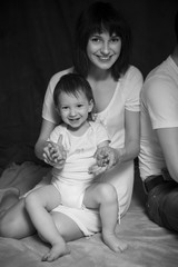 Little cute 2 years girl laying on mothers arms smiling. Neutral black background, black and white picture. All in white clothes. Happy family