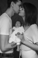 Little infant baby boy sleeping laying on mothers arms. Mother kissing father. Neutral black background, black and white picture. All in white clothes. Happy family