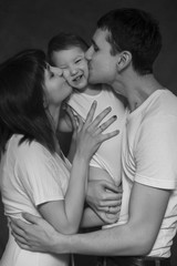 Little cute 2 years girl laying on mothers and fathers arms smiling, they are kissing her. Neutral black background, black and white picture. All in white clothes. Happy family
