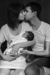 Little infant baby boy sleeping laying on mothers arms. Mother kissing father. Neutral black background, black and white picture. All in white clothes. Happy family