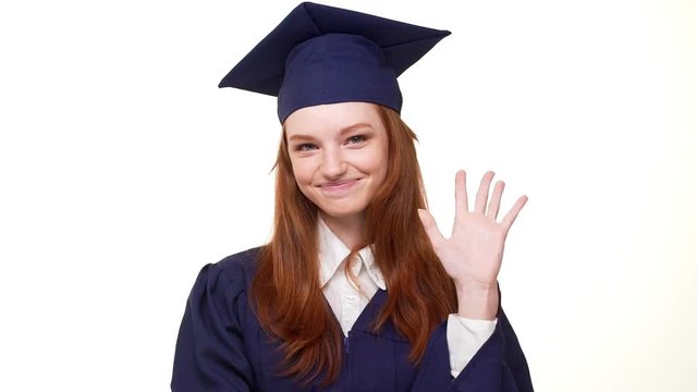 Beautiful young ginger Caucasian girl in blue academical dress standing smiling on white background with folded hands then waving arm. Footage in slowmotion