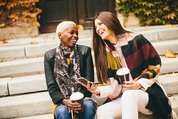 Multiracial friends sitting in autumn park, talking and carry coffee to go in the hands