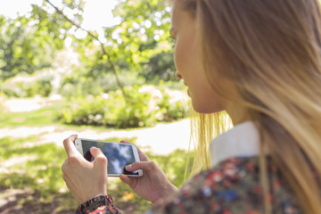 Beautiful brunette woman in neutral casual outfit walking in park, texting sms using her phone, smiling. Lifestyle portrait. Copy space