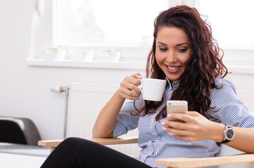 Concentrated young businesswoman drinking coffee and using mobile phone in home