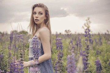 Beautiful brunette fit slim fragile woman with clear flawless skin and waved hair in sensual summer dress in the middle of meadow lupins field on a sunset, looking thoughtful and sad