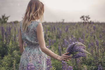 Beautiful brunette fit slim fragile woman with clear flawless skin and waved hair in sensual summer dress in the middle of meadow lupins field on a sunset, looking thoughtful and sad