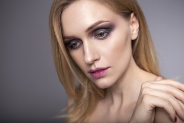 Fototapeta premium Blonde pretty woman in her 30th with fresh clean skin, smiling with smokey eyes and red lips make up on a neutral grey background. Casual posing