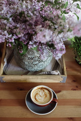 Beautiful blossoming purple lilac in a vase and fresh cappuccino in a cup with a saucer on the wooden table background, home set