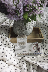 beautiful blossoming lilac in a tray with a glass of water, notebook and glasses in bed