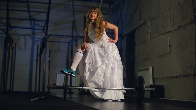 Beautiful and muscular bride blonde in a wedding dress doing an exercise with a barbell in a gym, crossfit