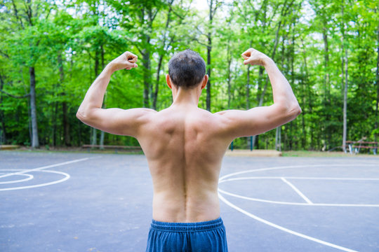 Back Of Young Fit Shirtless Man Flexing Muscles In Outdoors Outside Park During Summer