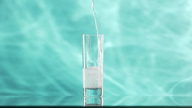 Clear sparkling water is pouring into collins glass standing on cyan smoky background in slowmotion. Water is filling glass in three quaters