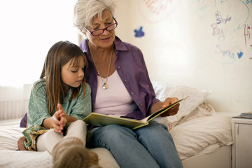 Grandmother reads a book with her young granddaughter as they sit together on a bed.