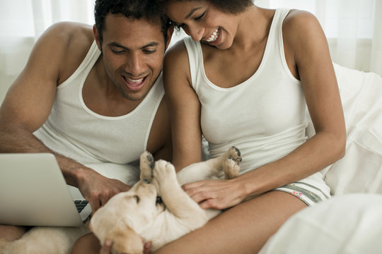 Couple tickling Labrador puppy while sitting on their bed.