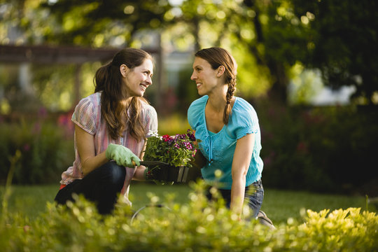 Two women smile and talk as they sit and kneel on the ground ready to plant flowers.