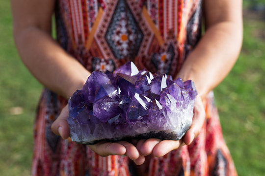 A young woman holds a large cluster of amethyst crystals as they shimmer in the sunlight.
