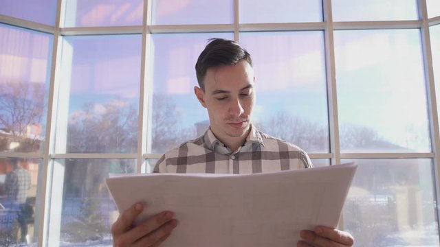 man looking drawings Stands on background of window in office. Attentive male with brown hair, wearing checkered shirt, standing Back to glass taking large sheets, analyzes images, graphics on paper