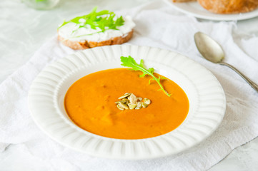 Vegan Creamy Delicious Pumkin Soup with fresh arugula and pamkin seeds and sliced bread on the white table. Selective focus.
