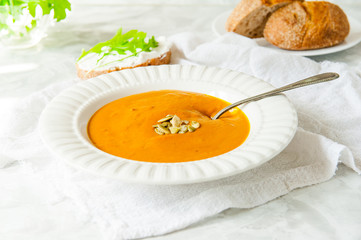 Vegan Creamy Delicious Pumkin Soup with fresh arugula and pamkin seeds and sliced bread on the white table. Selective focus.