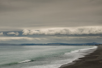 Napier, New Zealand - March 9, 2017: South side of Hawkes Bay with black lava beach and surf in front, all under heavy, dark storm clouds cut by band of white clouds. Shine on Pacific Ocean water.