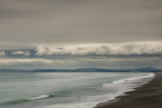 Napier, New Zealand - March 9, 2017: South side of Hawkes Bay with black lava beach and surf in front, all under heavy, dark storm clouds cut by band of white clouds. Shine on Pacific Ocean water.