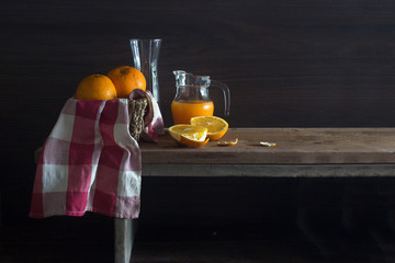 Fresh Oranges in rattan basket and glass with juice in jug on wooden table in dim light / Still...