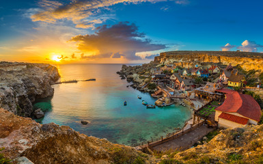 Il-Mellieha, Malta - Panoramic skyline view of the famous Popeye Village at Anchor Bay at sunset with traditional Luzzu boats, beautiful colorful clouds and sky - obrazy, fototapety, plakaty
