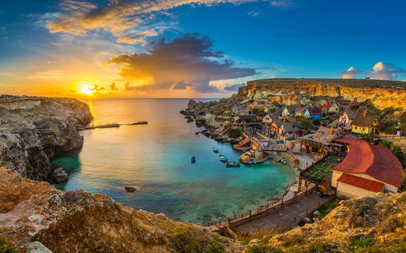Il-Mellieha, Malta - Panoramic skyline view of the famous Popeye Village at Anchor Bay at sunset with traditional Luzzu boats, beautiful colorful clouds and sky