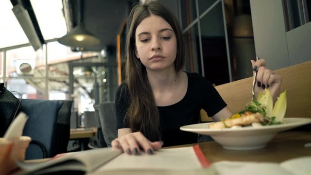 Teenage girl doing homework, reading textbook during meal in cafe

