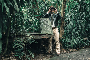 Explorer looking with binoculars standing in bush next to car wreckage.