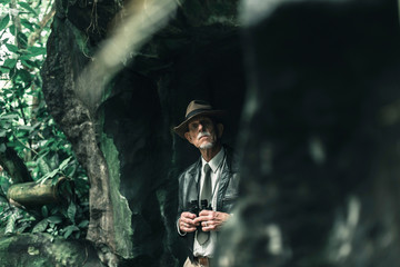 Explorer standing with binoculars in cave looking into distance.