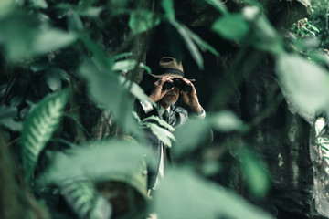 Man with hat and binoculars inspecting bush.