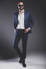 portrait of handsome fashion stylish hipster businessman model dressed in elegant blue suit in sunglasses posing on gray background in studio. Full length