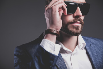 Obraz premium portret przystojny moda model stylowy hipster biznesmen ubrany w elegancki niebieski garnitur pozowanie na szarym tle w studio. Dotykając jego okularów przeciwsłonecznych