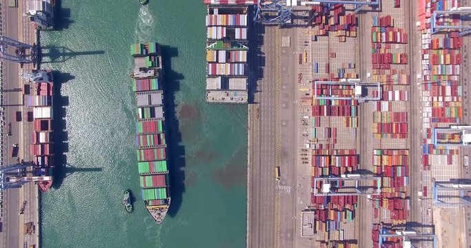 Commercial port with container ships during loading and unloading, and a container ship pulled by tugboats - Top down aerial view.