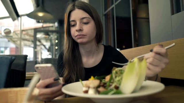 Young, teenage girl texting on smartphone and eating salad in cafe 
