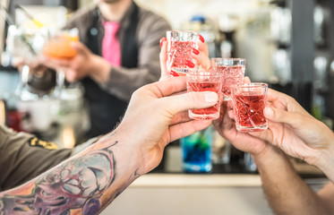 Group of drunk friends toasting cocktails at bar restautant - Food and beverage concept on nightlife moments - Defocused bartender serving drinks on background - Focus on hands cheering red shot glass - obrazy, fototapety, plakaty