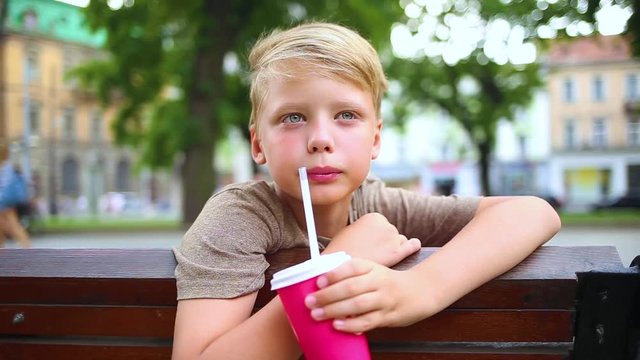 Cute white boy with blond hair drinks cold milkshake from red paper glass outdoors in summer park. Boy sits on wooden banch at walking blurry people background. Real time full hd video footage.