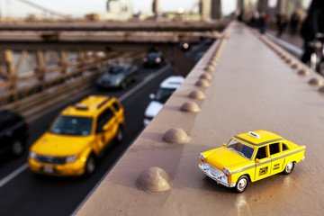 Model of a classic yellow taxi on a steel beam on Brooklyn Bridge, with a real taxi passing on the...