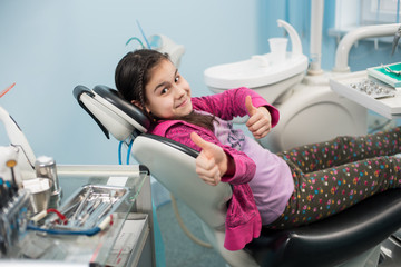 Happy patient girl showing thumbs up at dental office. Medicine, stomatology and health care concept. Dental equipment