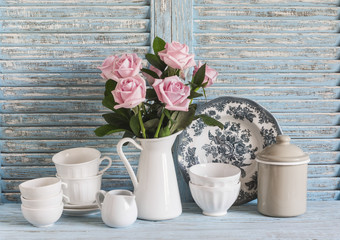 Fototapeta na wymiar Pink roses in a white enameled pitcher, vintage crockery on blue wooden rustic background. Kitchen still life in vintage style. Flat lay