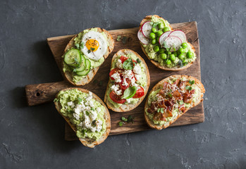 Breakfast avocado sandwich - with crispy bacon, quail egg, tomatoes, goat cheese, green peas, radish, cucumber on rustic cutting board. Healthy snack. On a dark background, top view
