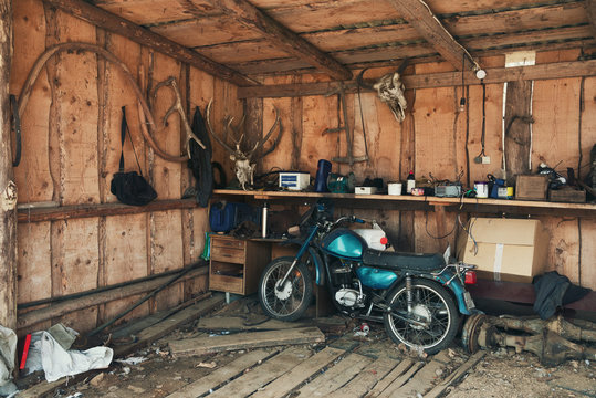 Old Blue-Green Motorbike In Picturesque Barn.Vintage Motorcycle In Old Hangar Against A Wall With Deer Antlers, A Bison Head And Many Interesting Rare Objects. Old Barn With Old Moped And Wooden Walls
