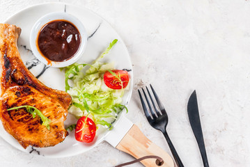 Healthy food. Portion of fresh salad with tomatoes, arugula, lettuce leaves, cabbage and grilled pork or veal cutlet on bone, barbecue sauce. On white marble plate, white table. Copy space top view