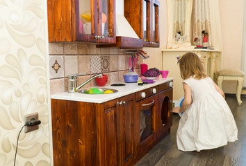 Active little preschool age child, cute toddler girl with blonde curly hair, shows playing kitchen, made of wood, plays in the kitchen