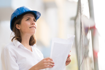 Cute female architect with blue hardhat checking office blueprints looking at something above her among scaffolding on construction site. Selective focus
