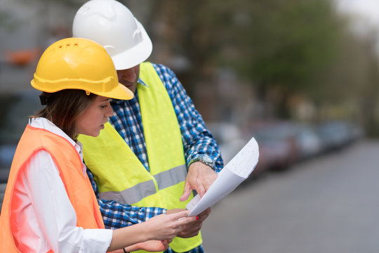 Male and female civil engineers wearing protective vests and hardhats checking office blueprints on construction site. Outdoors