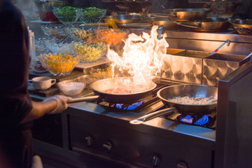 Chef in restaurant kitchen at stove with pan, doing flambe on food. low ligth selective focus.