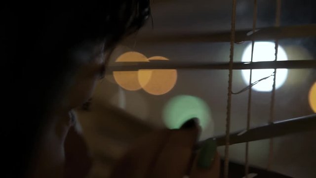 Young brunette girl looks through the window through the blinds to the street, lit by street lamps, pulls out the curtains and leaves, close-up, view of the city from the side. Reflection in the glass